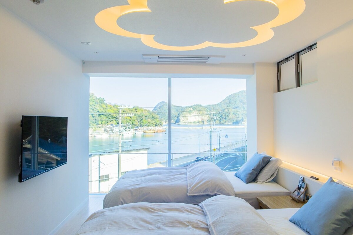 A hotel room with a bed, a sofa, a TV, and a view of a body of water and hills.
