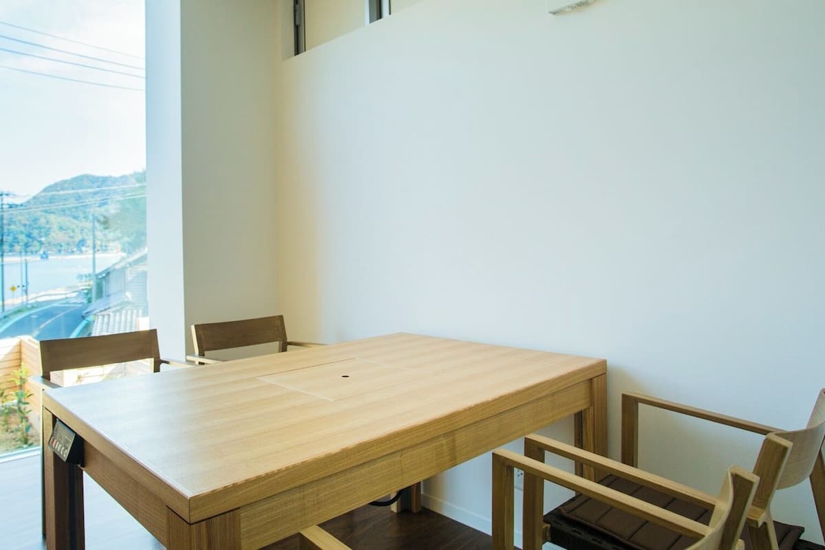 Un comedor con mesa y sillas de madera, vista a un cuerpo de agua y colinas, y una ventana con cables de electricidad.