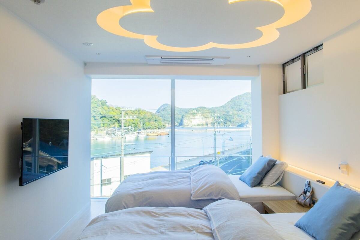 Habitación de hotel con cama, sofá, televisor y vistas al agua y colinas.