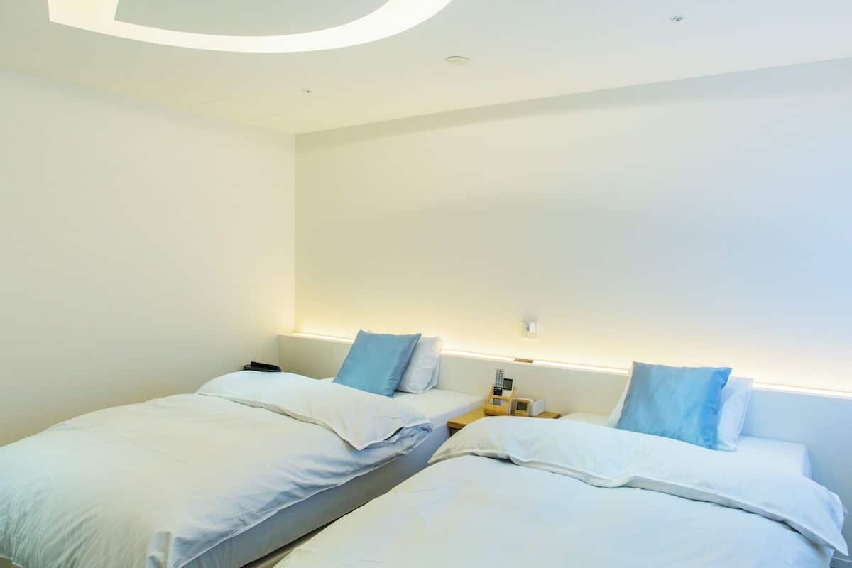 A modern hotel room with two beds, a bedside table with a lamp, and a large window.