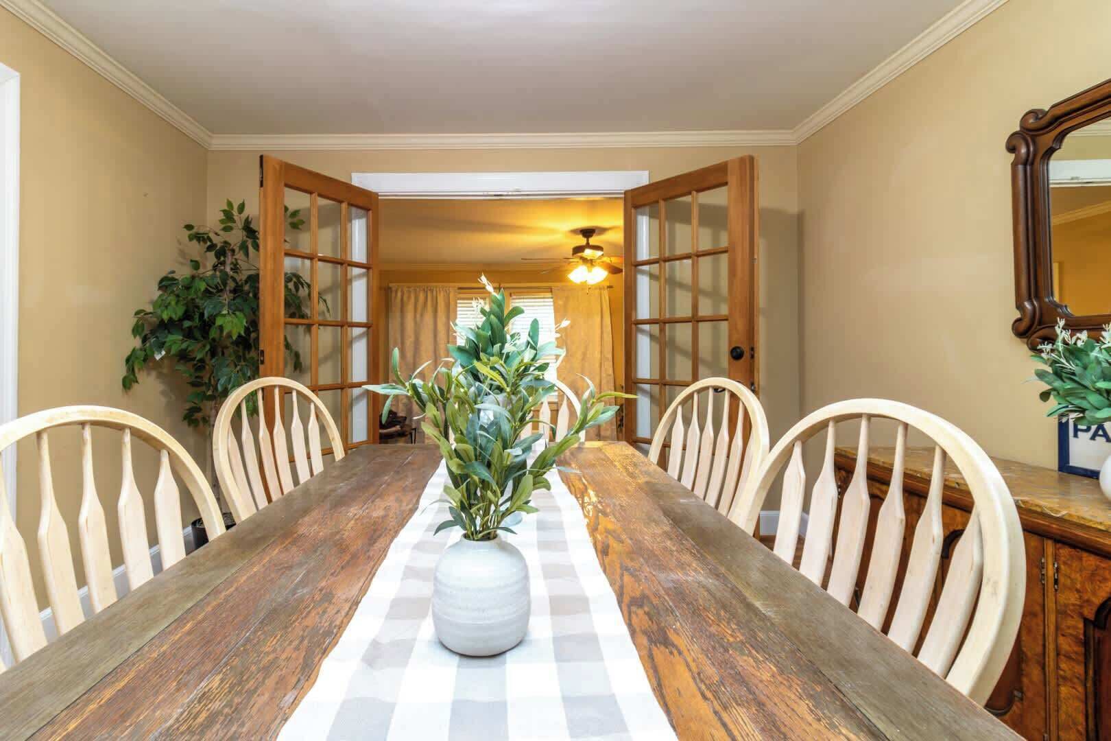 A dining room with a wooden table, white chairs, a vase with green plants, and a ceiling fan.