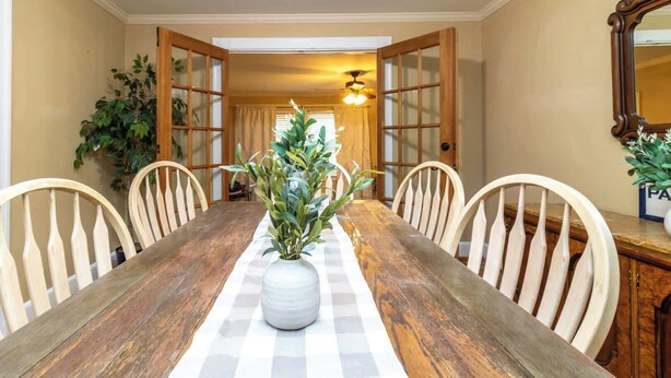 Een eetkamer met een houten tafel, witte stoelen, een vaas met groene planten en een plafondventilator.