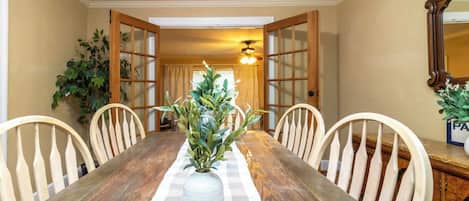 A dining room with a wooden table, white chairs, a vase with green plants, and a ceiling fan.