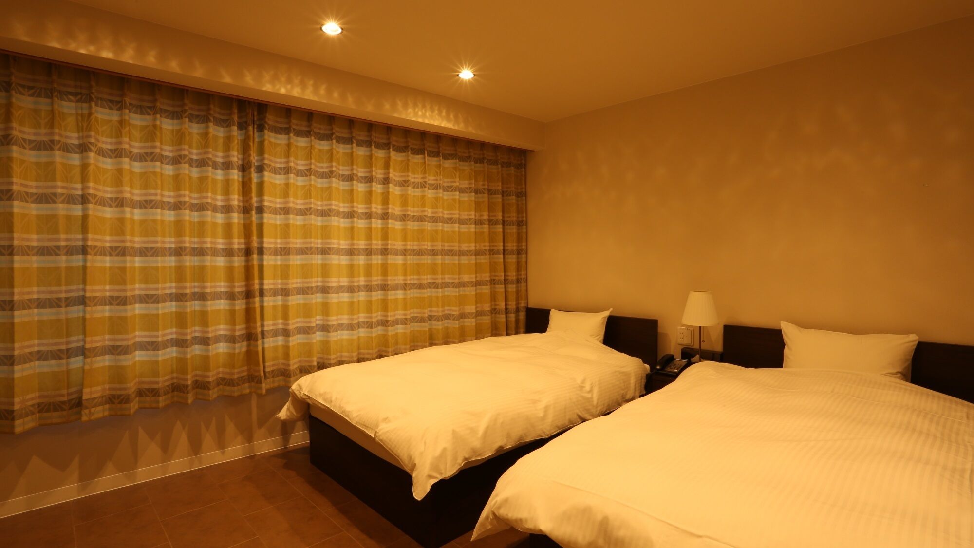 A hotel room with two beds, a large window with curtains, a bedside table with a lamp, and a phone.