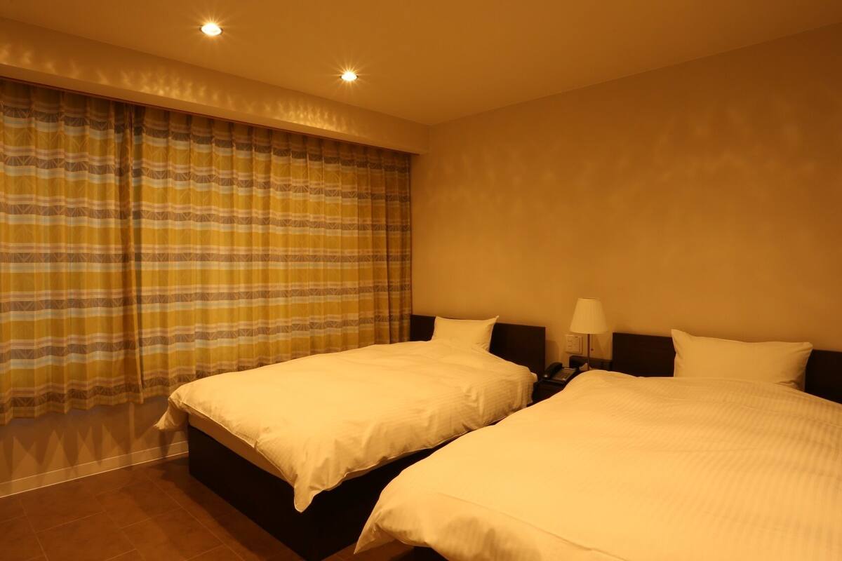 A hotel room with two beds, a large window with curtains, a bedside table with a lamp, and a phone.