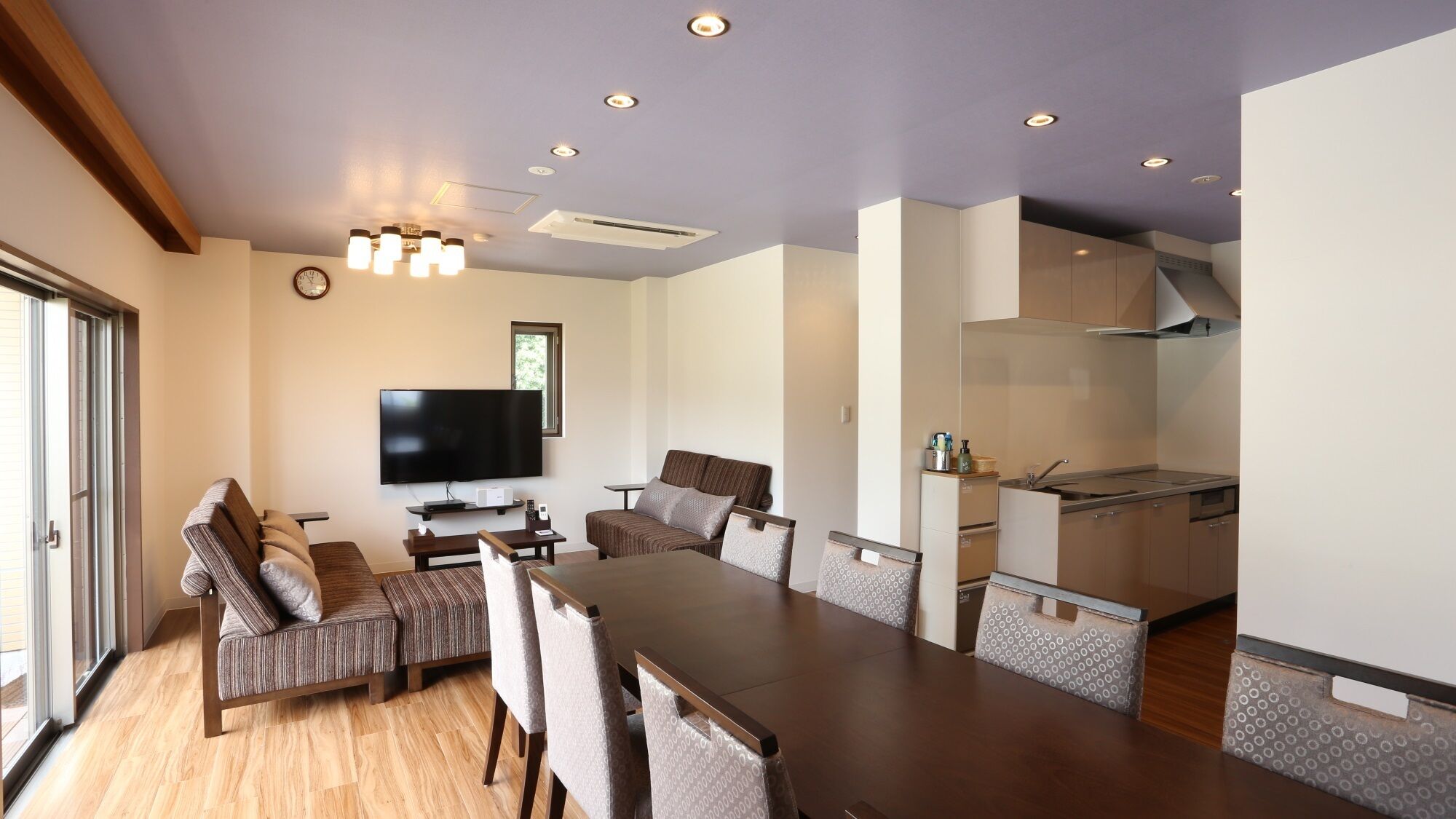 A modern living room with a flat-screen TV, a dining table, and a kitchen area with cabinets and a sink.