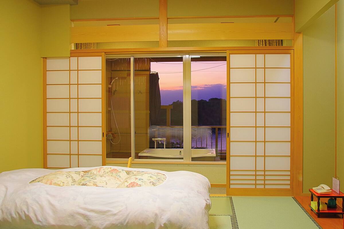 Una habitación tradicional japonesa con suelo de tatami, una cama con colcha floral, una puerta corredera de tipo shoji y vistas a la montaña al atardecer.