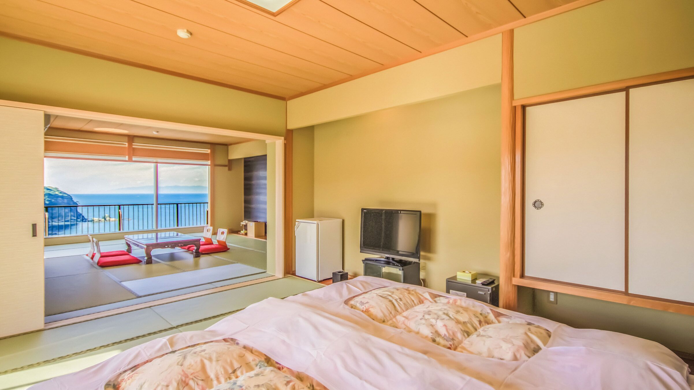 Een kamer met een bed, een televisie, een koelkast en een schuifdeur die leidt naar een balkon met uitzicht op zee.