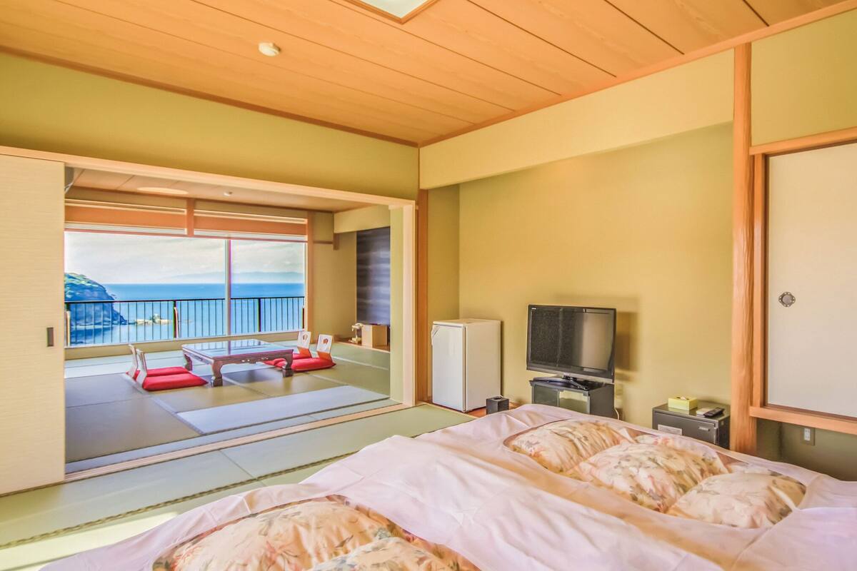 Una habitación con cama, televisión, refrigerador y puerta corrediza que da a un balcón con vista al mar.