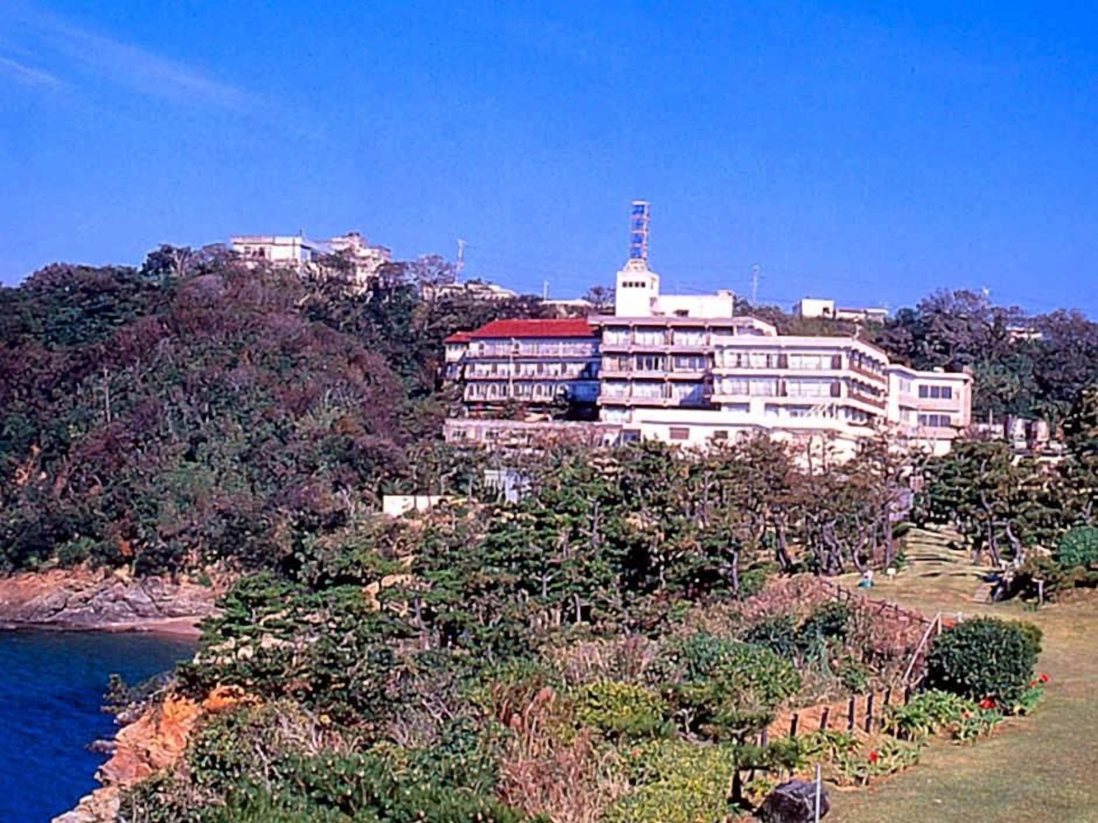 Um edifício de vários andares com varandas, localizado numa encosta coberta por vegetação densa e com um corpo de água ao primeiro plano.