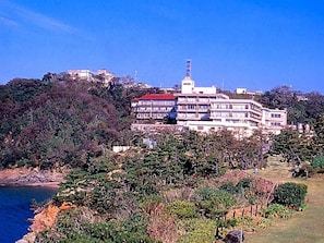 Un edificio de varios pisos con balcones, ubicado en una ladera con vegetación densa y un cuerpo de agua al frente.