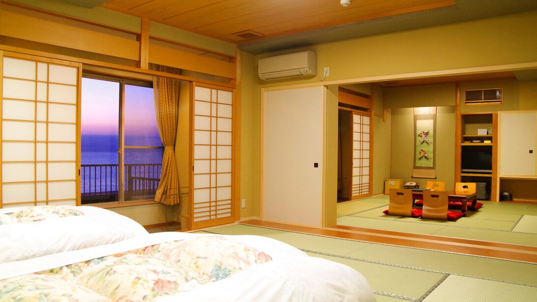 Une pièce de style japonais traditionnel, avec un sol recouvert de tatamis, des portes coulissantes, un lit avec une literie à motifs floraux, une télévision et une vue sur la mer.