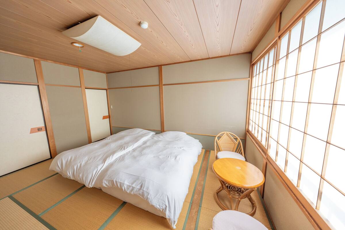 Una habitación japonesa tradicional con suelo de tatami, una cama con ropa de cama blanca, una pequeña mesa de madera y una ventana con diseño de cuadrícula.