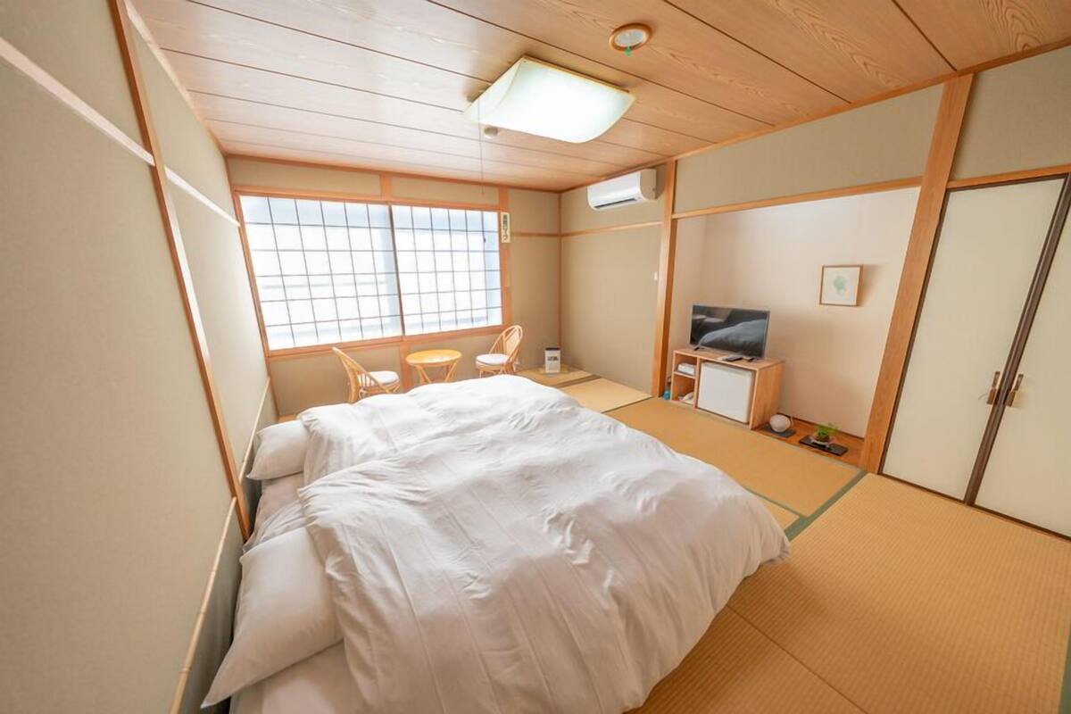 Una habitación tradicional japonesa con suelo de tatami, una cama con ropa blanca, un televisor, un escritorio pequeño y un armario.