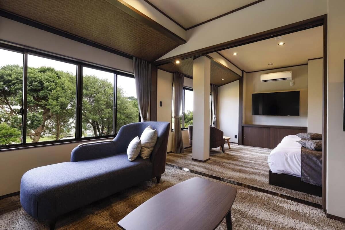 Habitación de hotel moderna con sofá, cama, televisor y vistas a árboles a través de amplias ventanas.