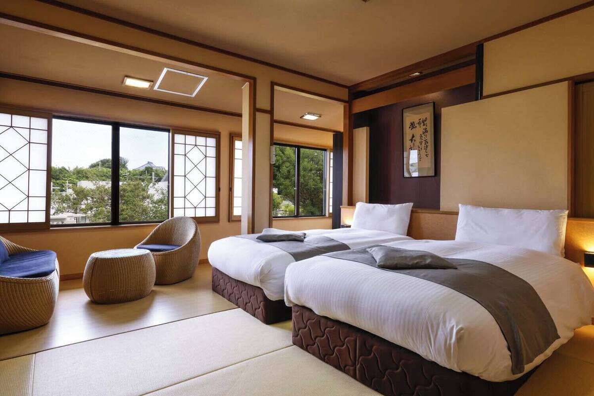 Habitación de hotel con suelo de tatami, una cama grande con cabecera, dos zonas de estar con cojines y amplias ventanas con vistas a zonas verdes.