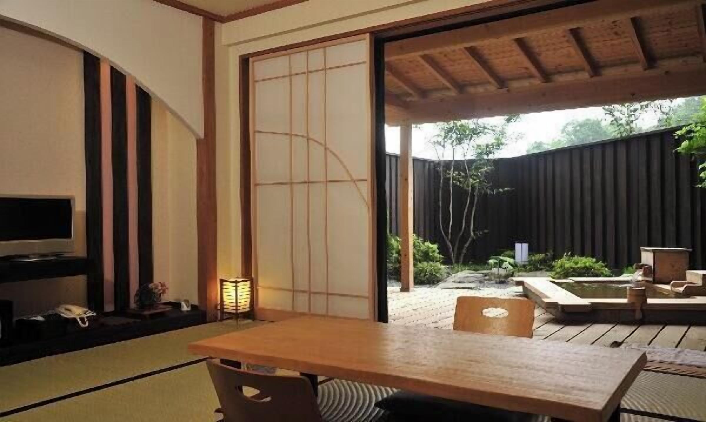 A traditional Japanese room with tatami flooring, a low table, a television, and a sliding door leading to an outdoor area with a wooden deck and a small water feature.