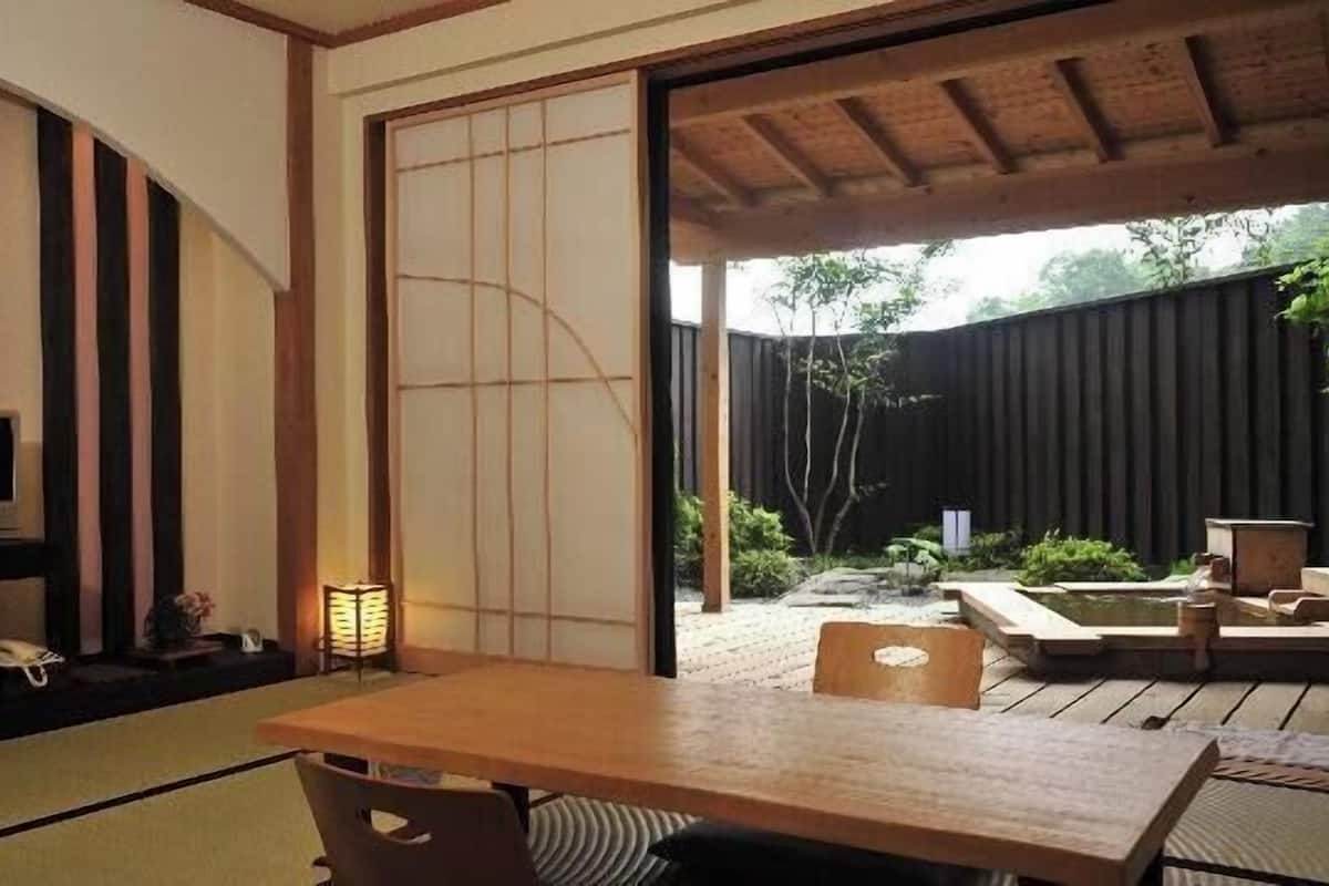 A traditional Japanese room with tatami flooring, a low table, a television, and a sliding door leading to an outdoor area with a wooden deck and a small water feature.