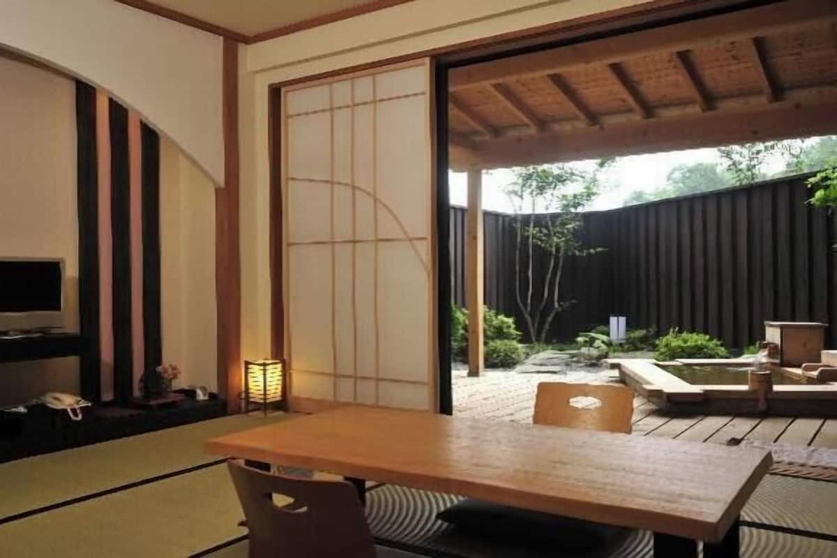 A traditional Japanese room with tatami flooring, a low table, a television, and a sliding door leading to an outdoor garden.