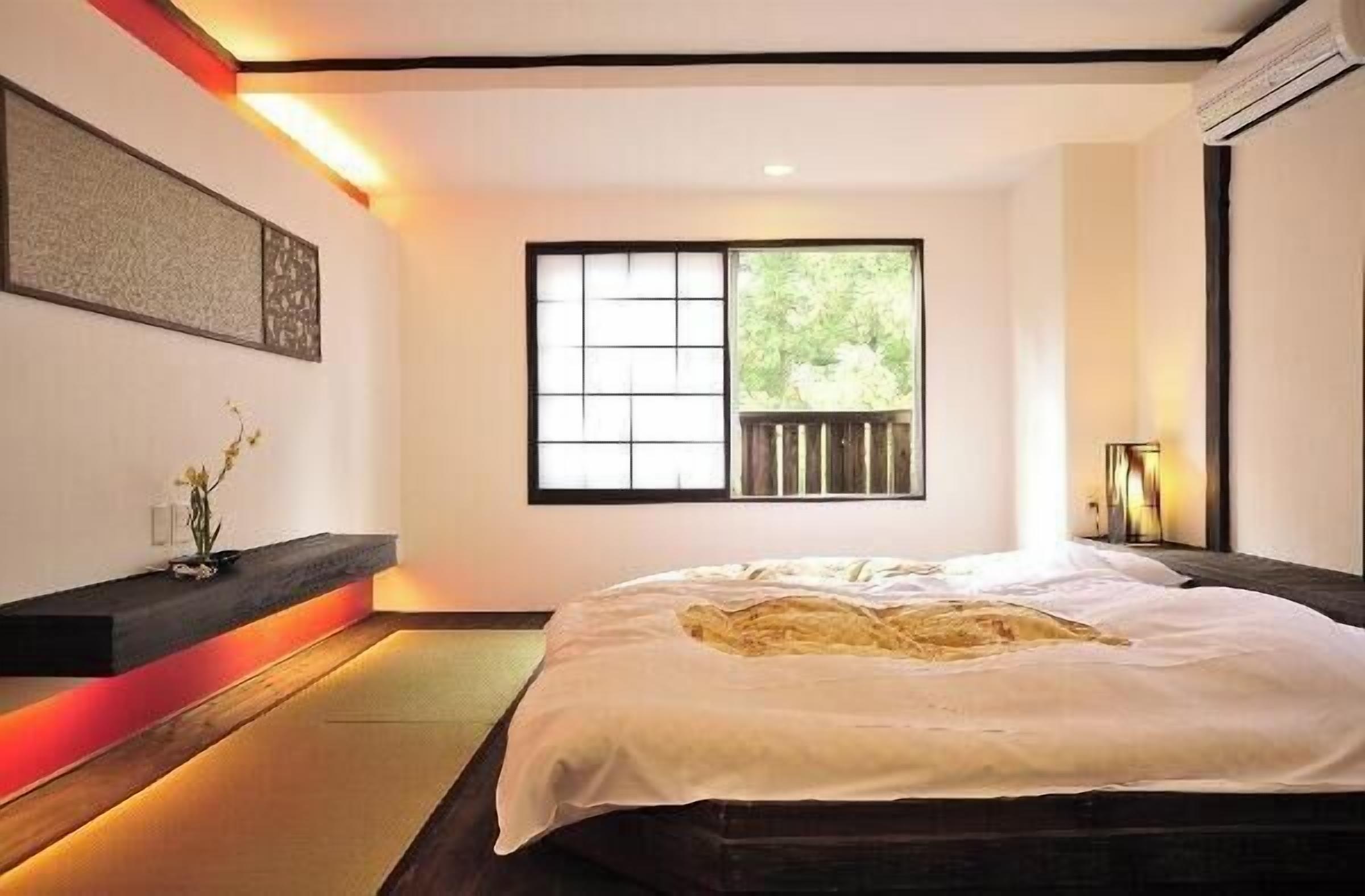 A bedroom with a bed, a window, a wall-mounted shelf with a vase, and a bedside table.