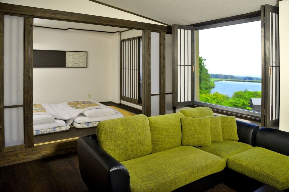A room with a green sofa, a bed with white bedding, a window with a view of a lake, and a wooden ceiling.