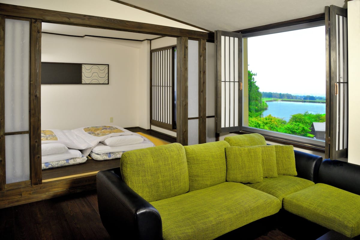 A room with a green sofa, a bed with white bedding, a window with a view of greenery and water, and a wooden ceiling structure.