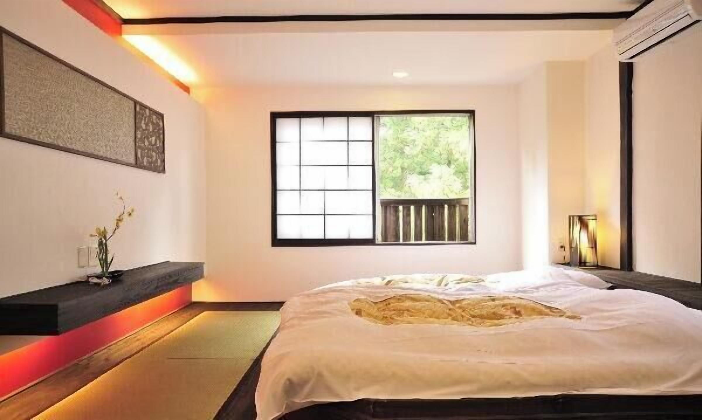 A minimalist bedroom with a large bed, a window with a view of greenery, a wall-mounted artwork, and a bedside table with a lamp.