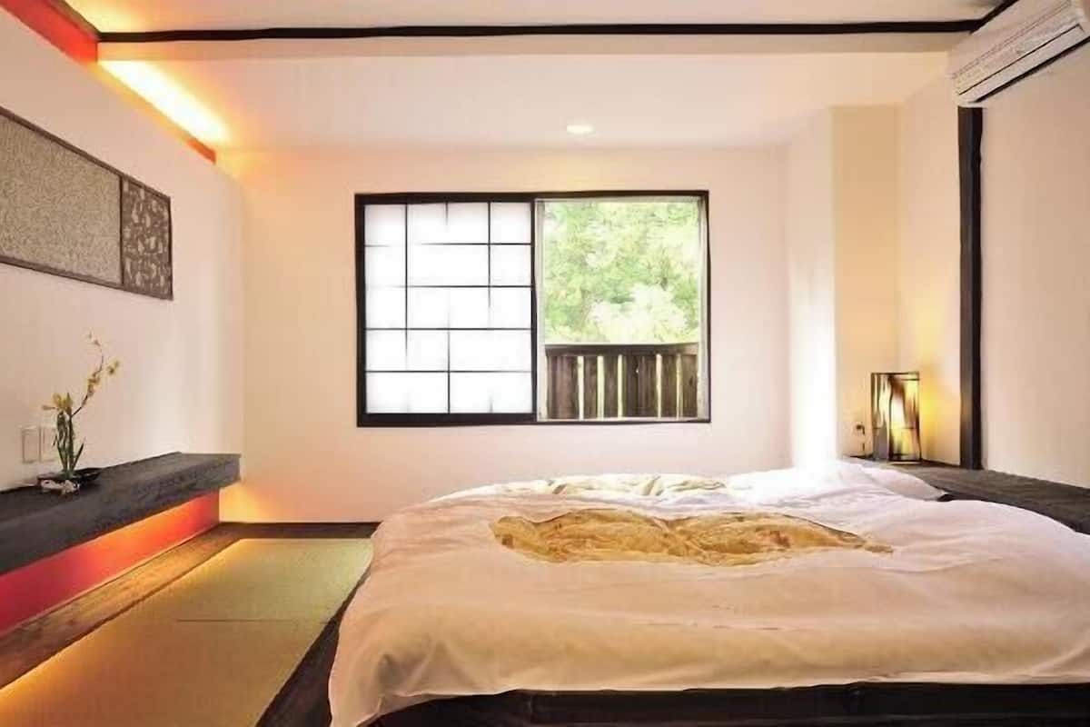 A minimalist bedroom with a large bed, a window with a view of greenery, a wall-mounted artwork, and a bedside table with a lamp.