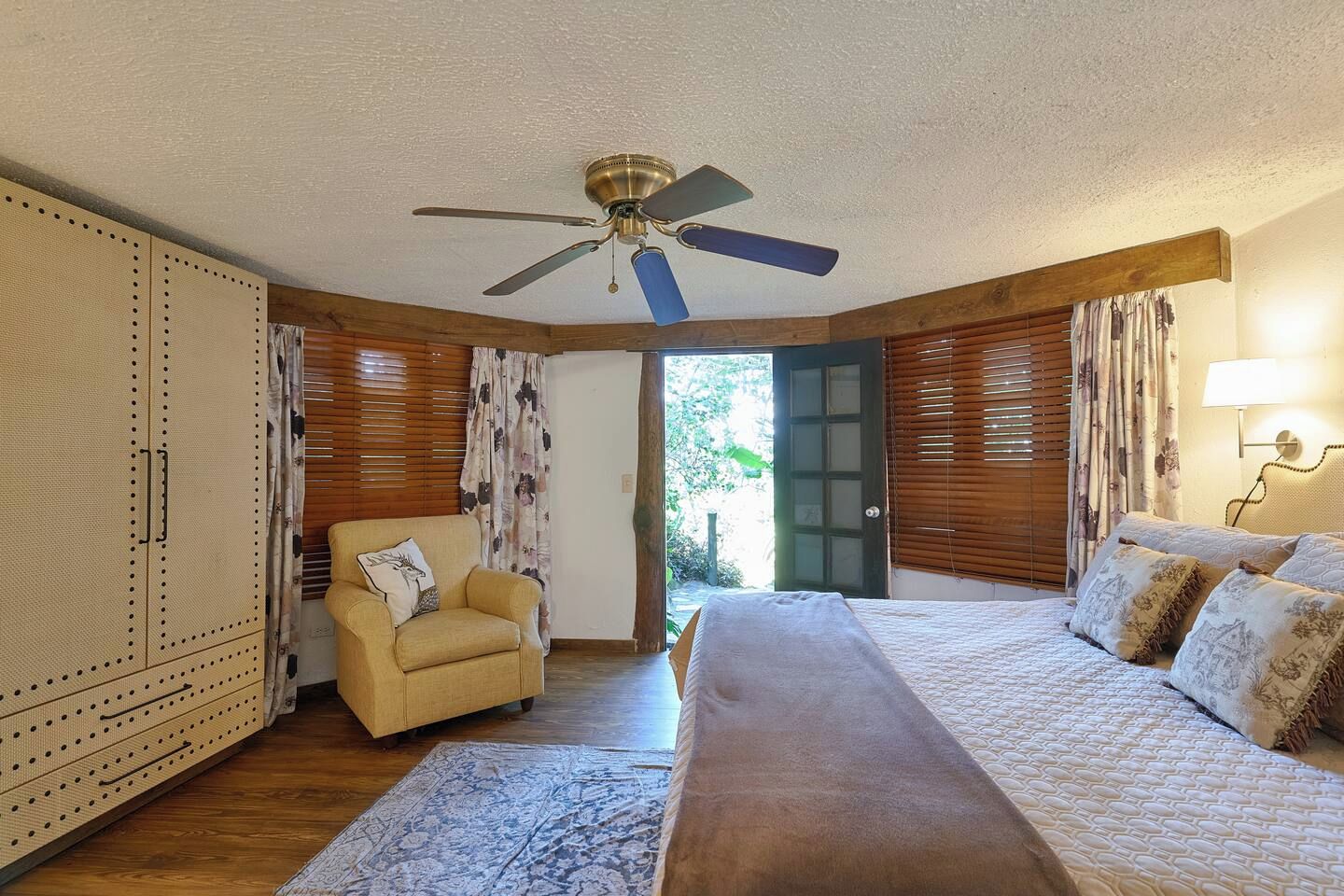 A bedroom with a bed, a chair, a ceiling fan, and a door leading outside.