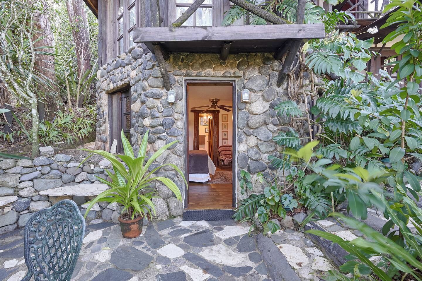 Une entrée en pierre menant à un bâtiment doté d’une avancée en bois, avec un pot de fleurs et une chaise placés à l’extérieur.