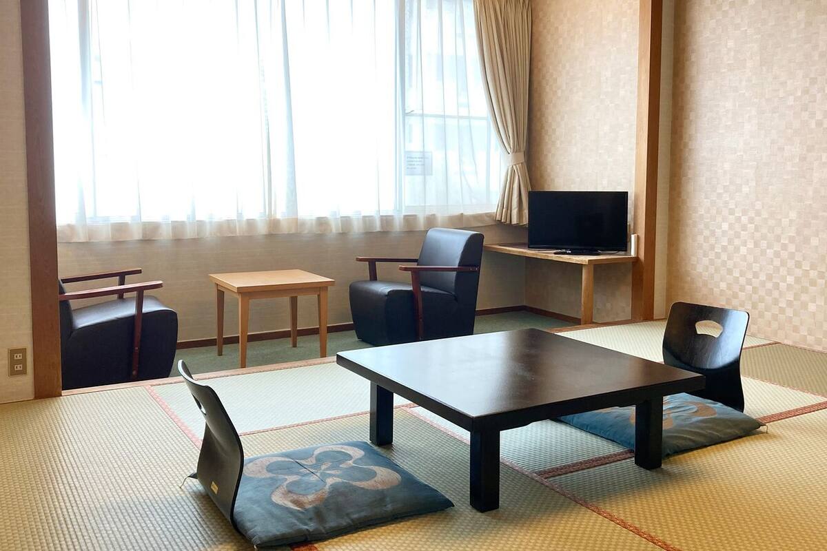 A traditional Japanese room with tatami flooring, a low table, chairs, a television, and a window with curtains.