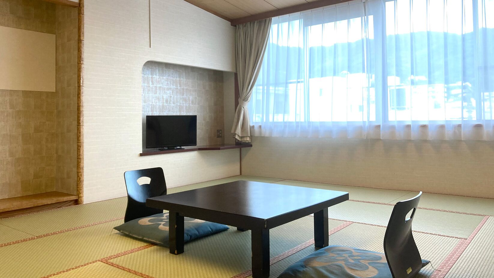 A traditional Japanese room with tatami flooring, a low table, two chairs, a television on a shelf, and a large window with sheer curtains.