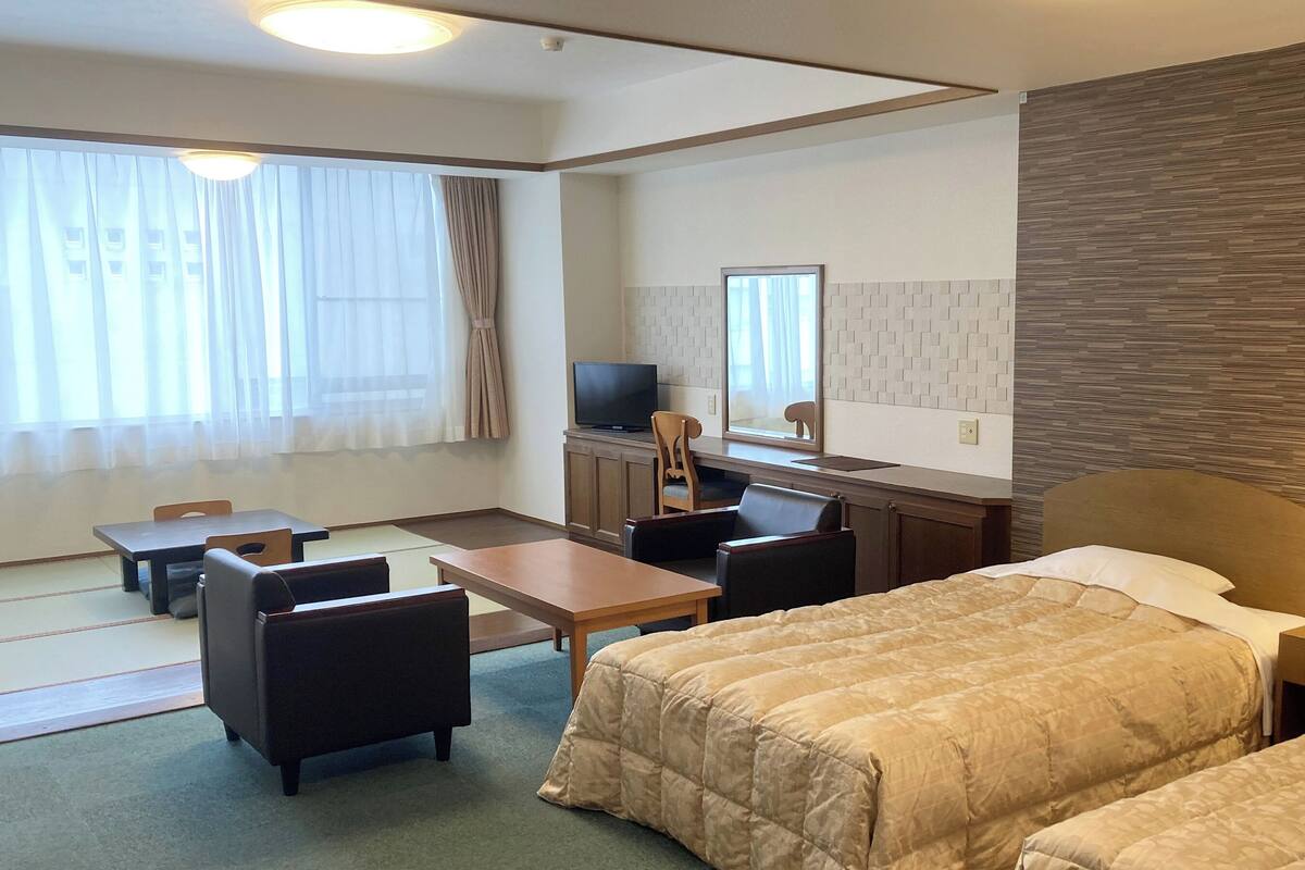 A hotel room with a bed, a desk with a chair, a TV, and a window with curtains.