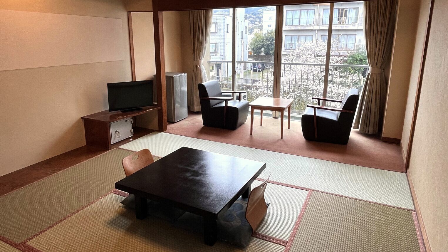 A room with tatami flooring, a television, a refrigerator, and a balcony with a view of buildings and cherry blossoms.