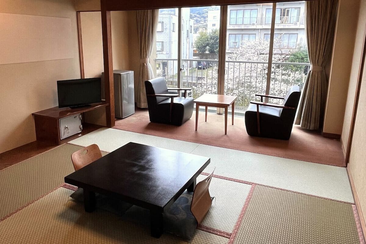 A room with tatami flooring, a television, a refrigerator, and a balcony with a view of buildings and cherry blossoms.