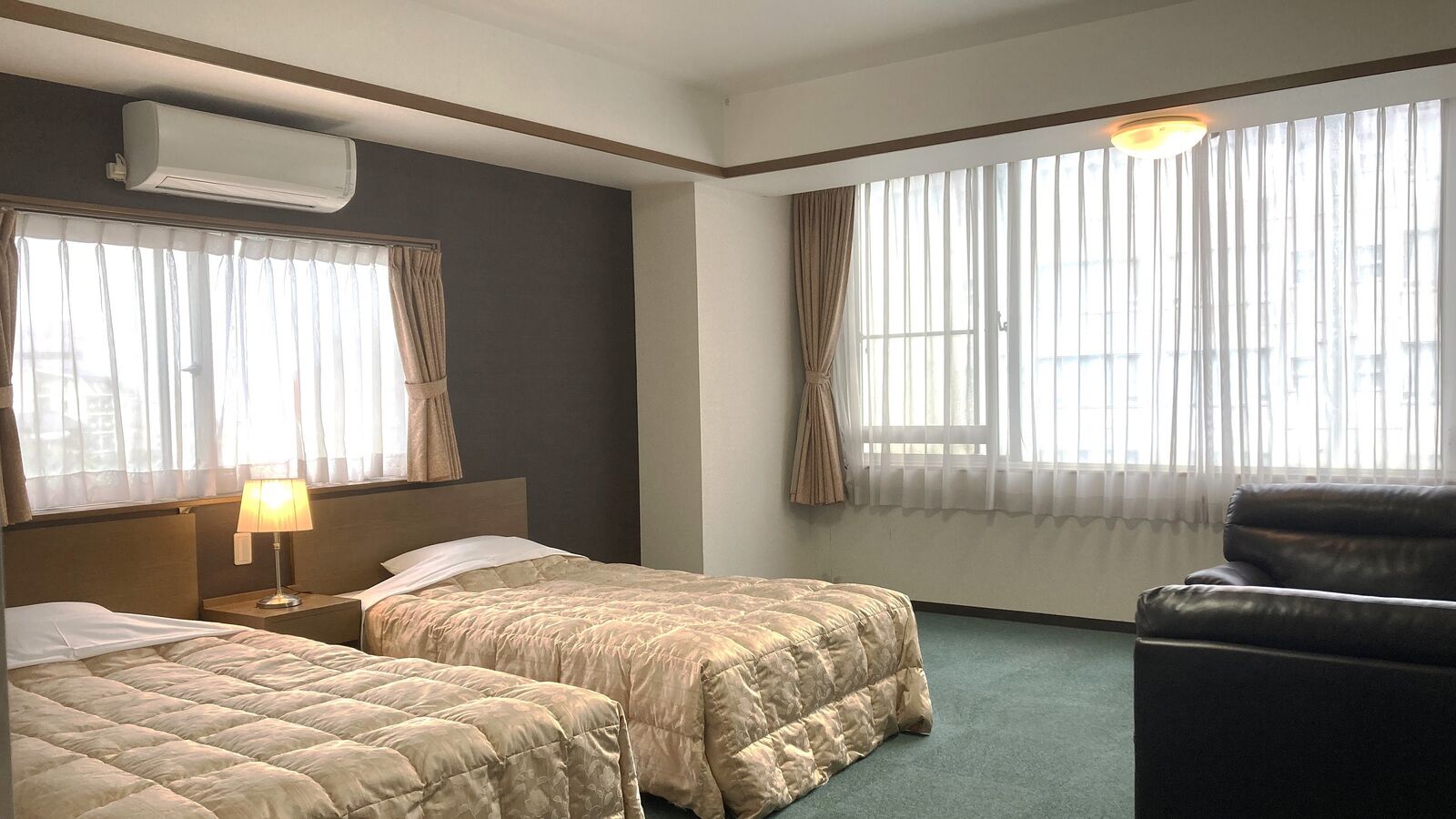 A hotel room with two beds, a desk with a lamp, a leather chair, and large windows with curtains.