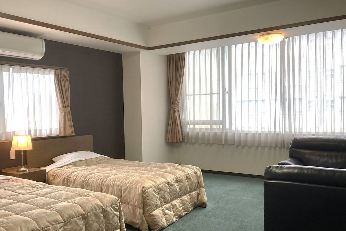 A hotel room with two beds, a desk with a lamp, a leather chair, and large windows with curtains.