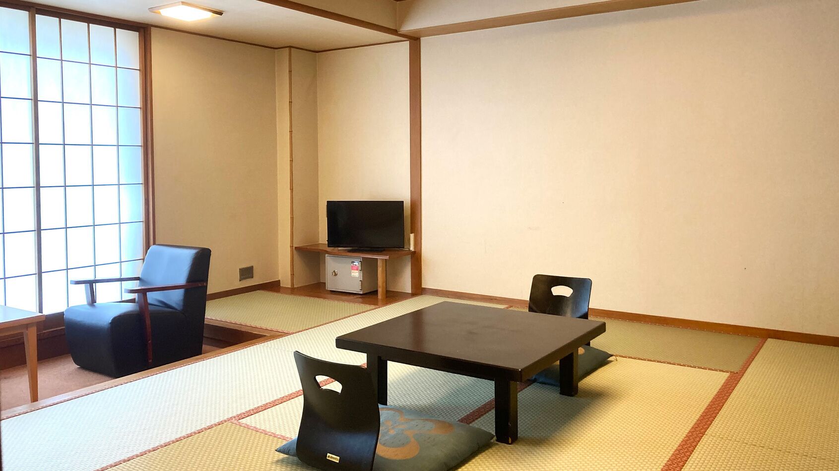A room with tatami flooring, a television on a stand, a table, and two chairs.