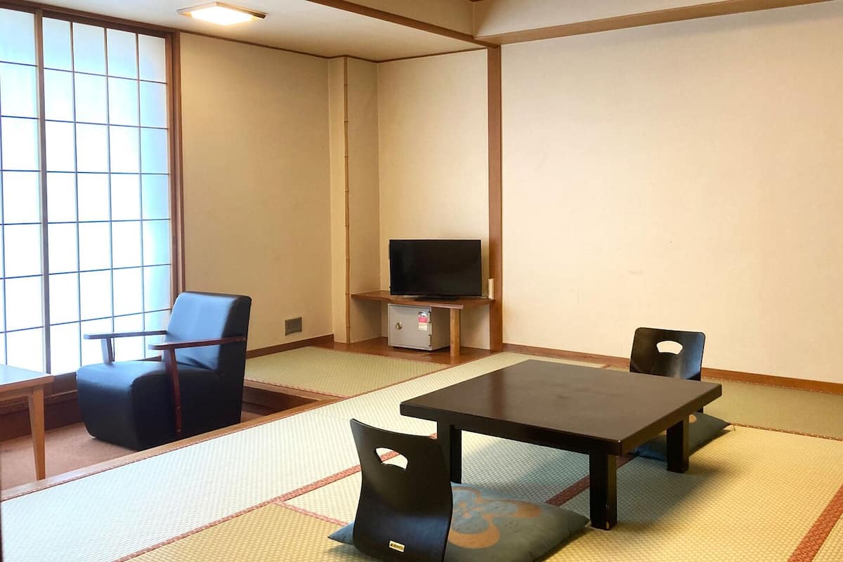 A room with tatami flooring, a television on a stand, a table, and two chairs.