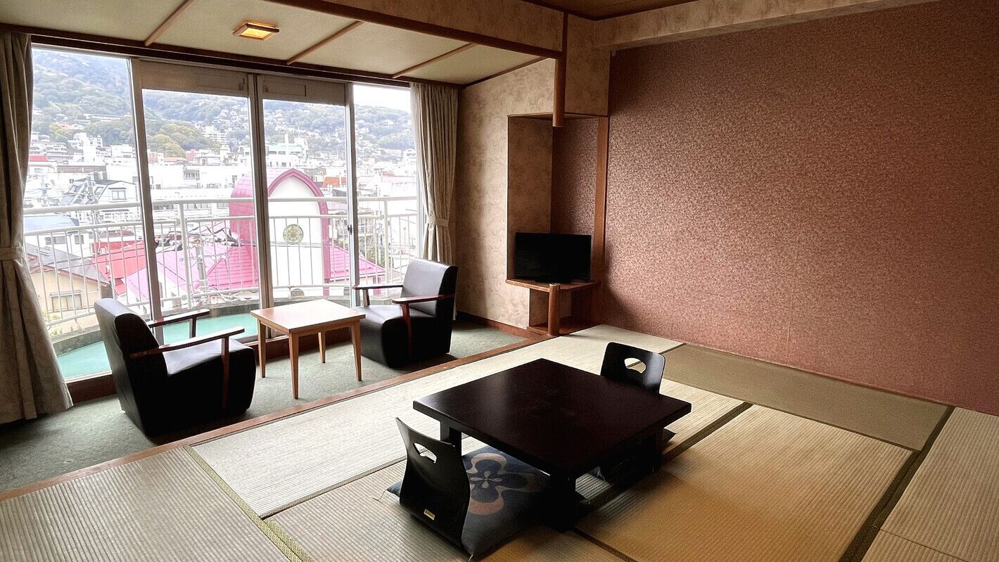 A room with tatami flooring, a dining table, chairs, a TV, and a large window with a view of buildings.