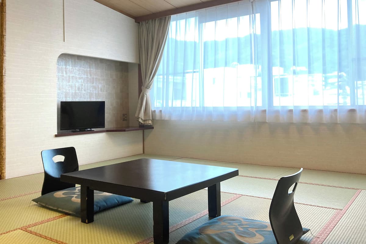 A room with tatami flooring, a low table, two chairs, a television on a shelf, and a large window with curtains.