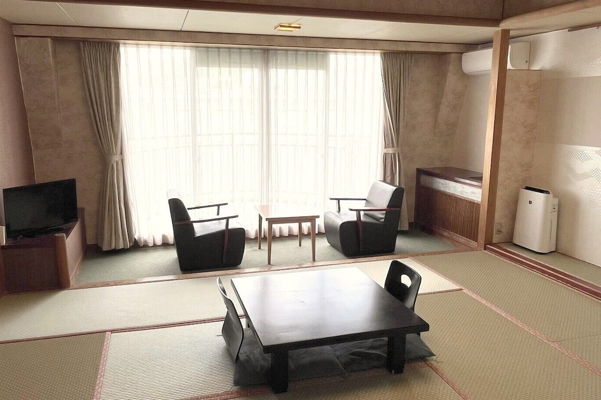 A room with tatami flooring, a table, two chairs, a television, and a radiator.