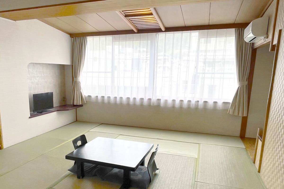 A room with tatami flooring, a low table, a chair, a TV on a shelf, and a large window with curtains.