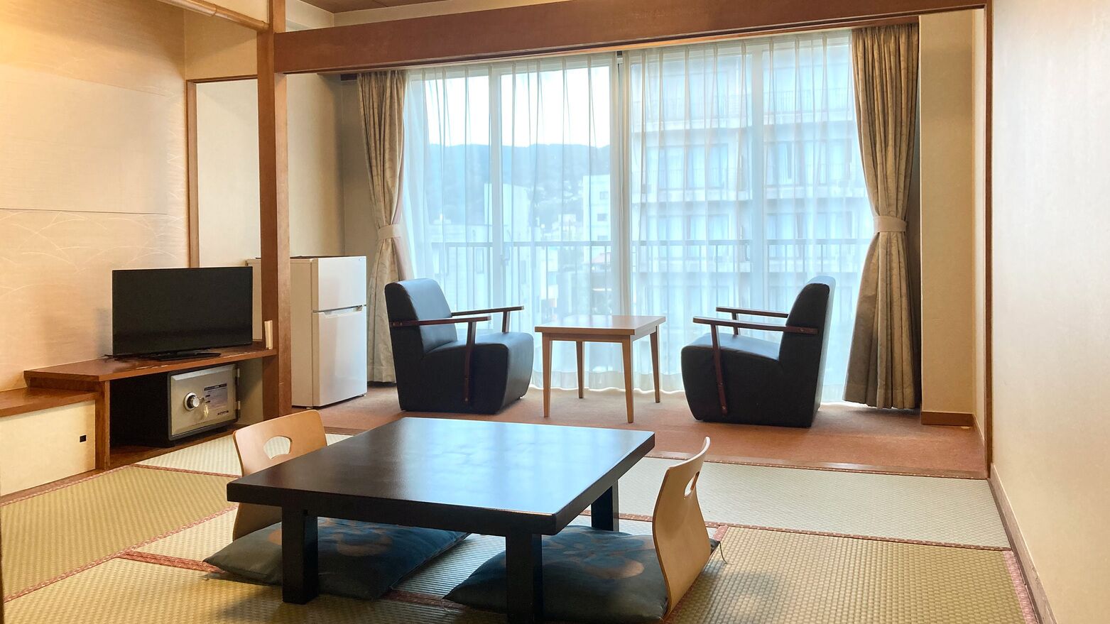 A hotel room with a TV, two chairs, a small table, and a view of buildings through large windows.