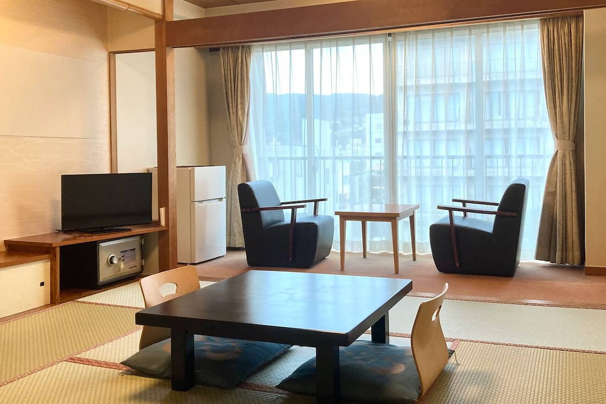 A hotel room with a TV, two chairs, a small table, and a view of buildings through large windows.