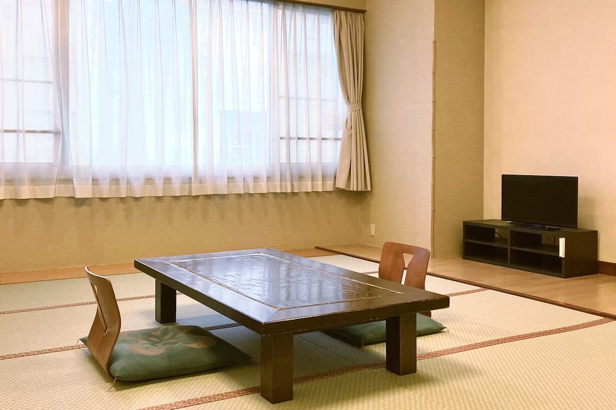 A room with tatami flooring, a low table with cushions, a television, and a window with sheer curtains.