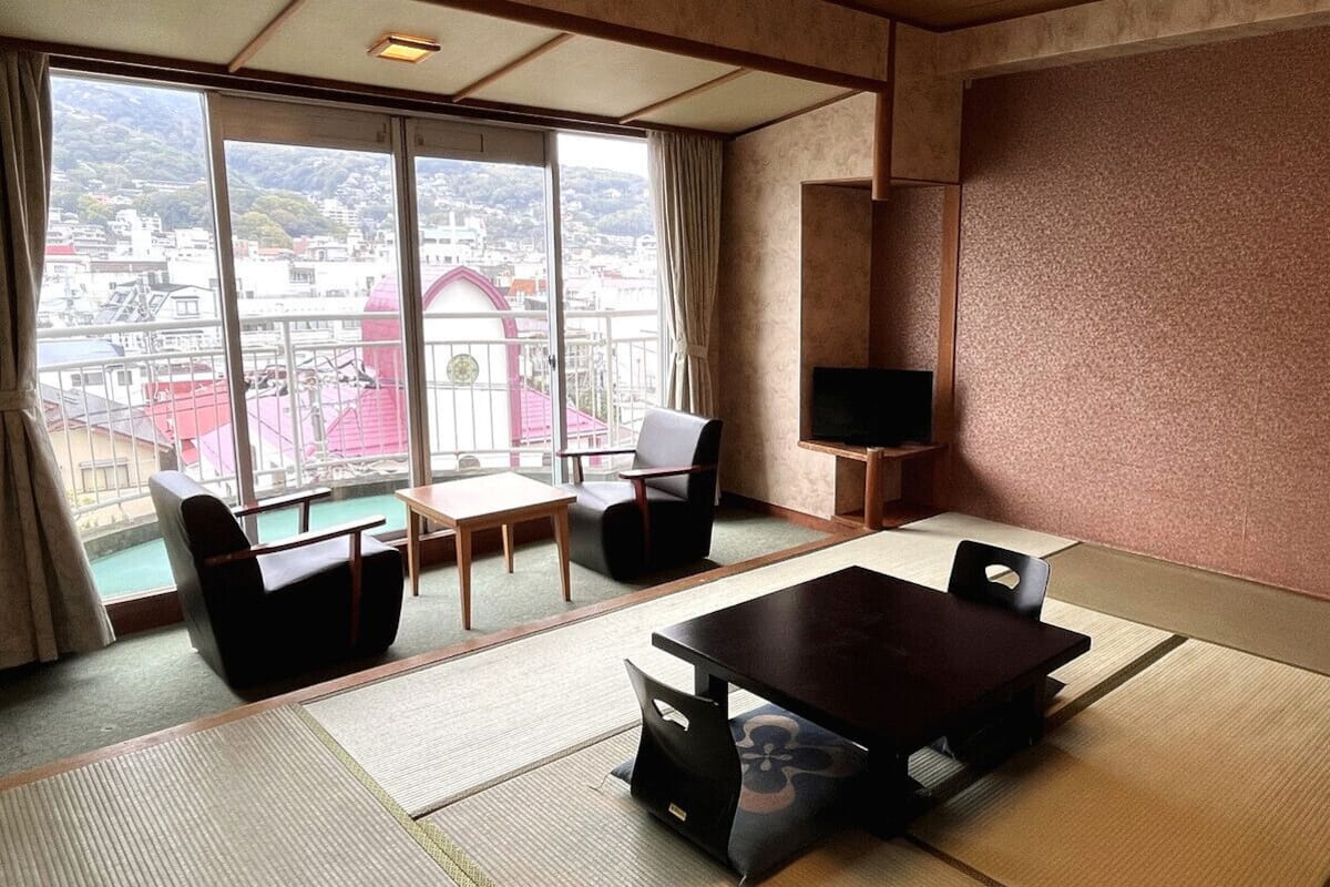 A room with tatami flooring, a dining table, chairs, a TV, and a large window with a view of buildings.