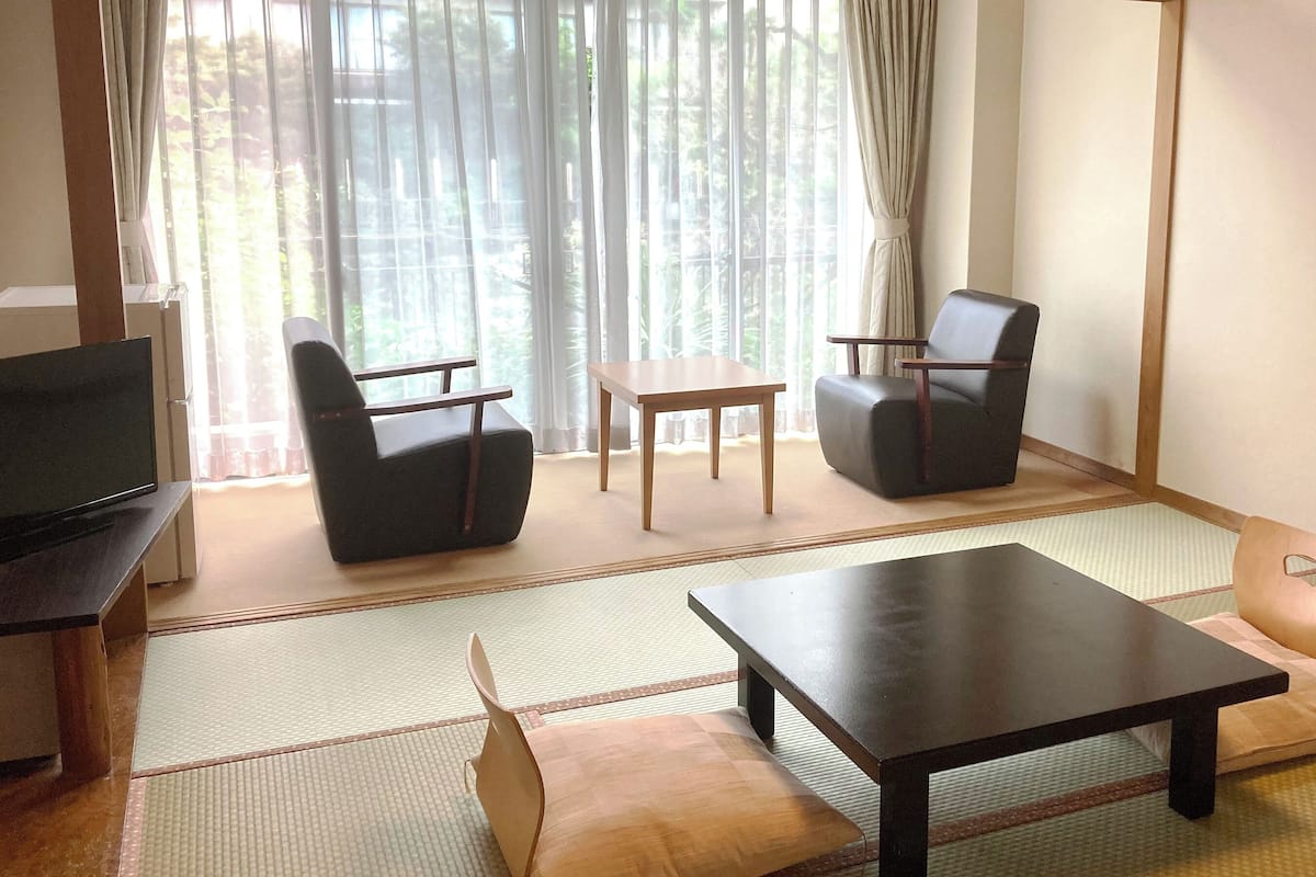 A room with tatami flooring, a television, two chairs, a small table, and a view through a window with curtains.