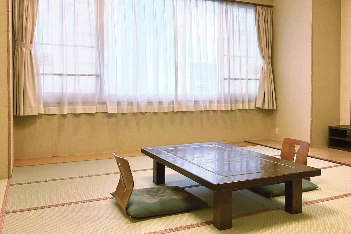 A room with tatami flooring, a low table with cushions, a television on a stand, and a window with sheer curtains.
