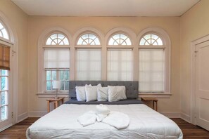 Un dormitorio con una cama grande, cabecera gris y tres ventanas arqueadas con persianas.